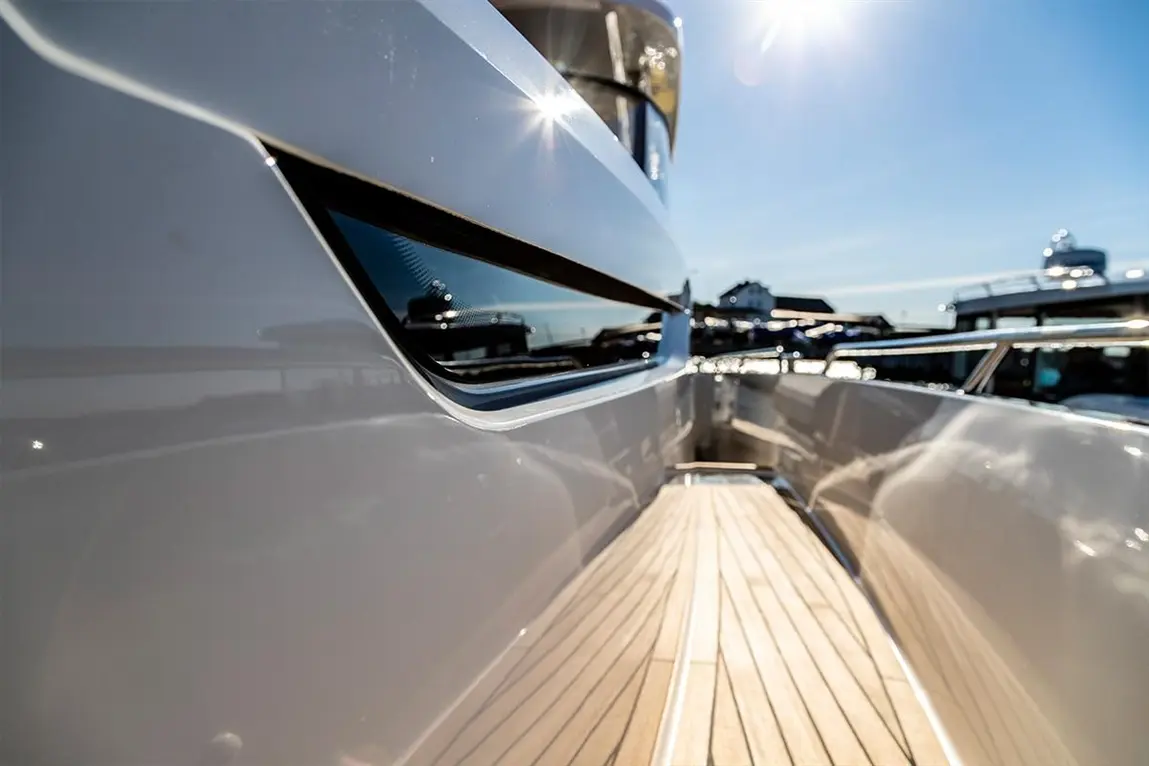 Close-up of a sleek yacht's side, featuring polished surfaces and a wooden deck, with boats in the background under a sunny sky.